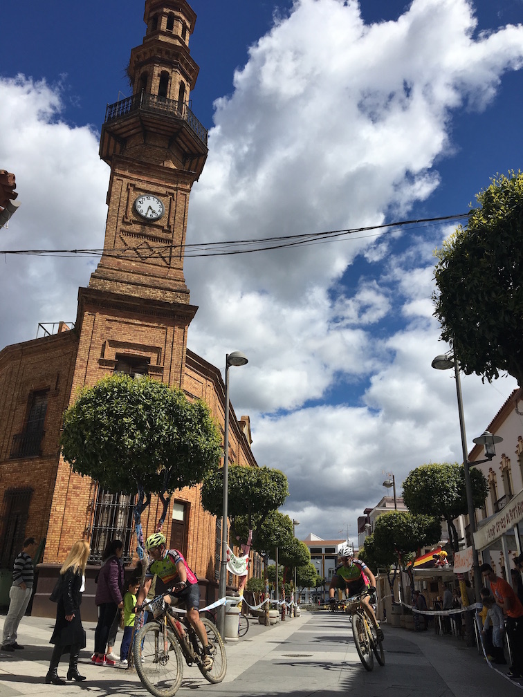 Los ciclistas a su paso por la Avenida de Andalucía en Nerva, junto a la torre del reloj del Ayuntamiento